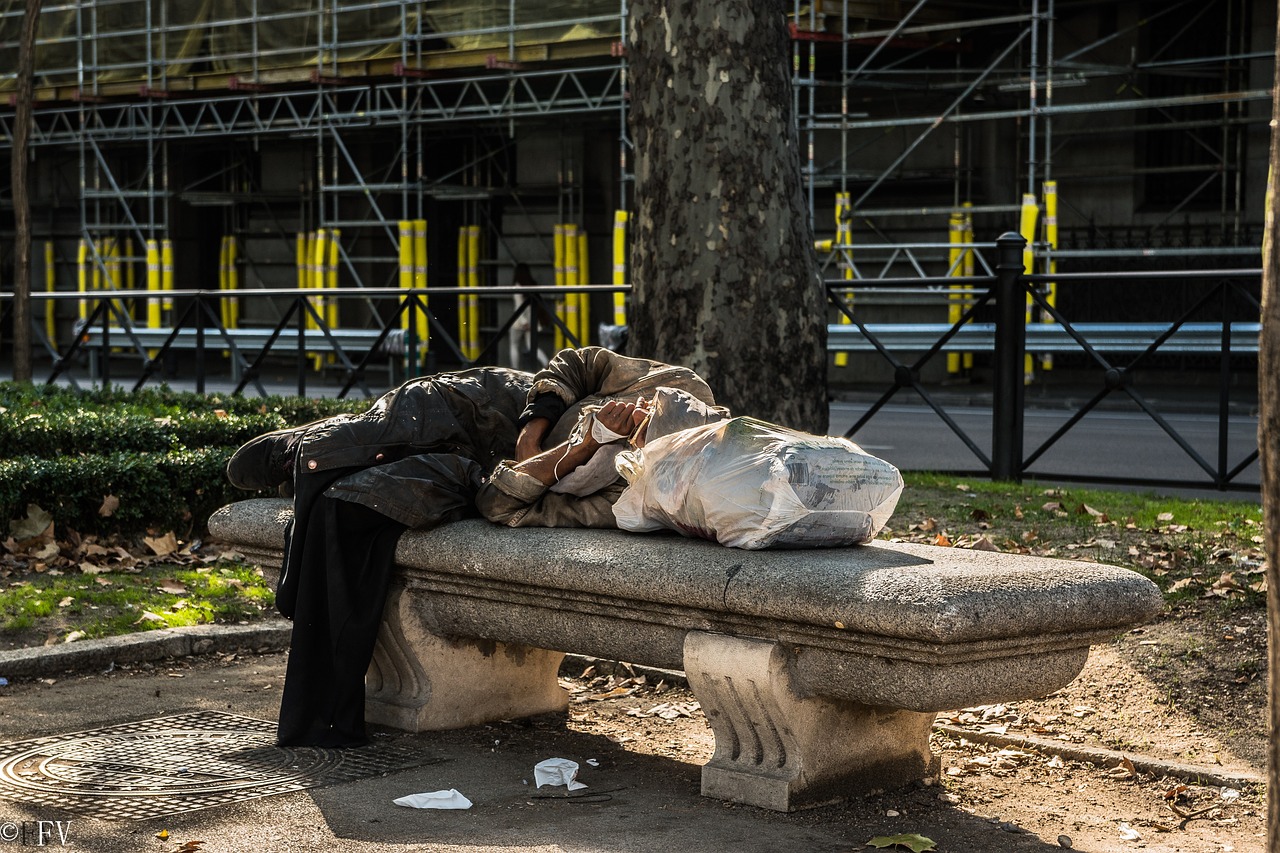 A homeless person sleeping on a stone bench in a city environment with scaffolding behind them.